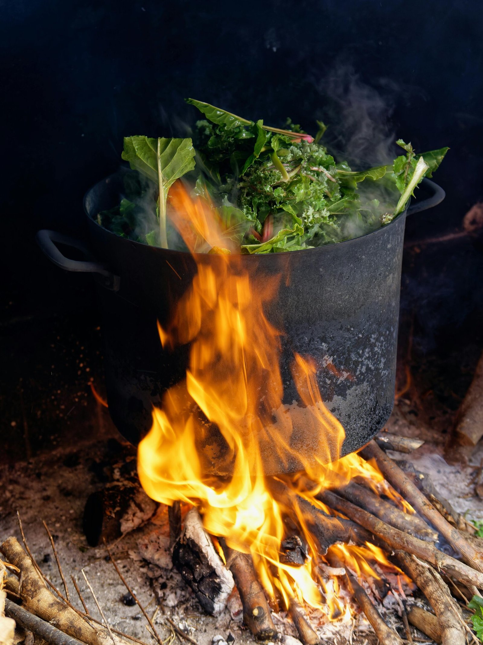 A pot with greens cooking over an open wood fire, emitting smoke and flames.